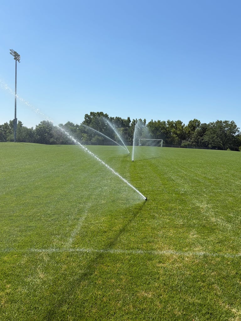 Soccer field with sprinklers watering grass, goal net visible in background under clear sky