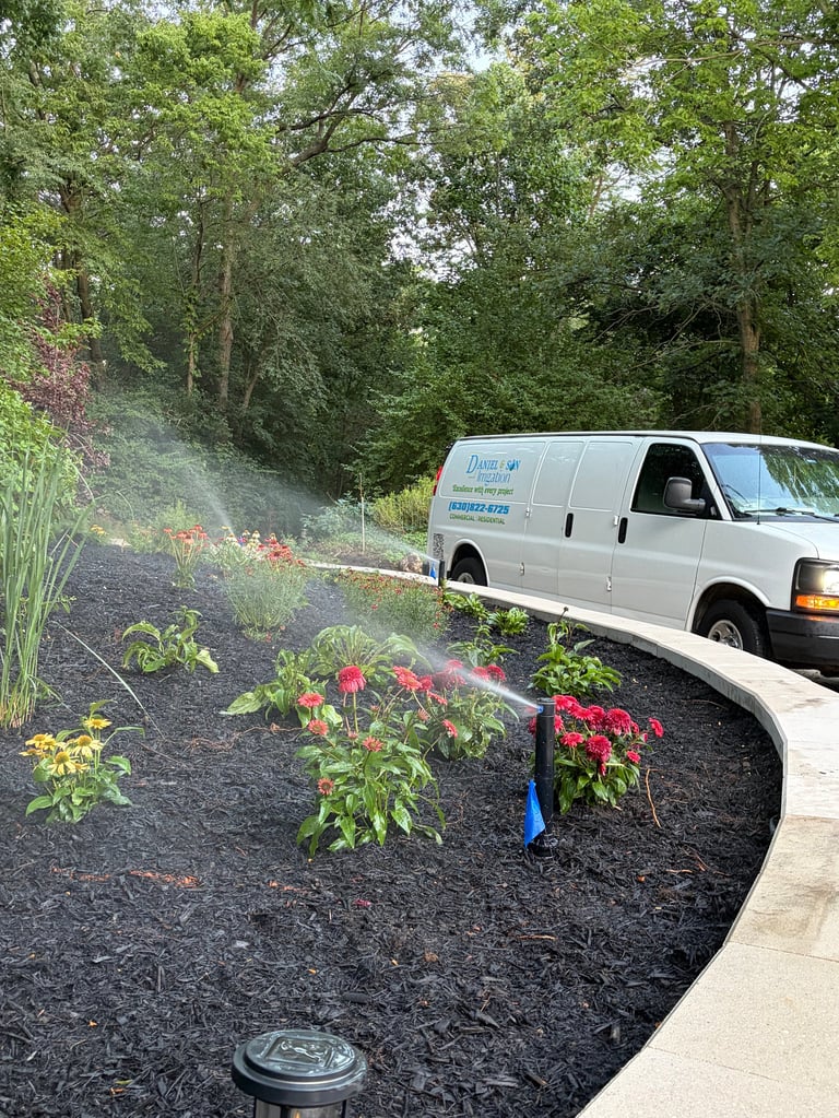 Landscaping service van spraying water on flower beds with red flowers and black mulch near trees